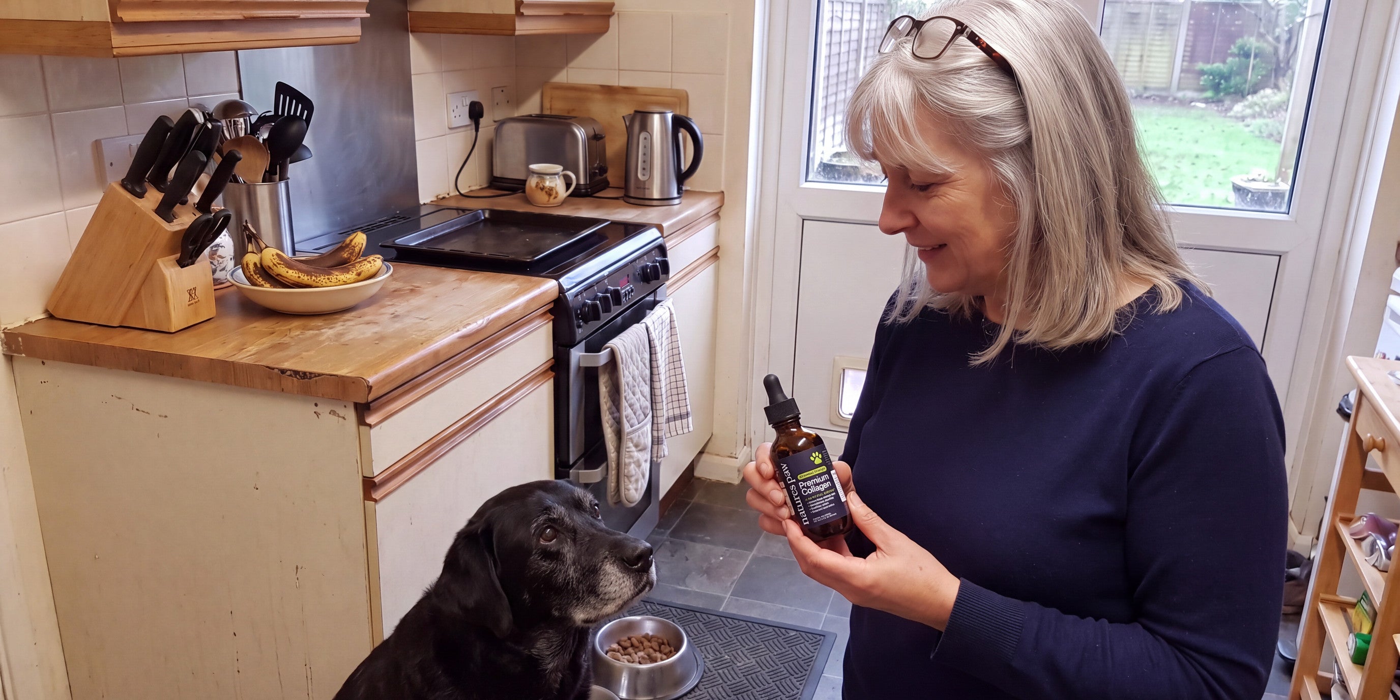 Woman in a kitchen holding the Nature's Paw bottle, an old black Labrador at her feet
